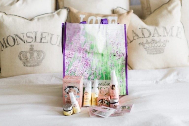 A collection of beauty and skincare products are displayed in front of a decorative bag with a lavender field design, reminiscent of the tranquility one might seek during a home birth. The setup includes various bottles, containers, and boxes. The background shows pillows with the words "MONSIEUR" and "MADAME.