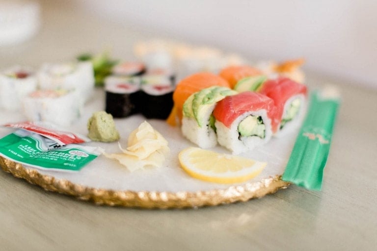 A plate of assorted sushi with various types of rolls including tuna, salmon, and avocado, along with pieces of pickled ginger, wasabi, and a lemon slice. A packet of soy sauce and a pair of green chopsticks are also on the plate—raising the question, can you eat sushi while pregnant?