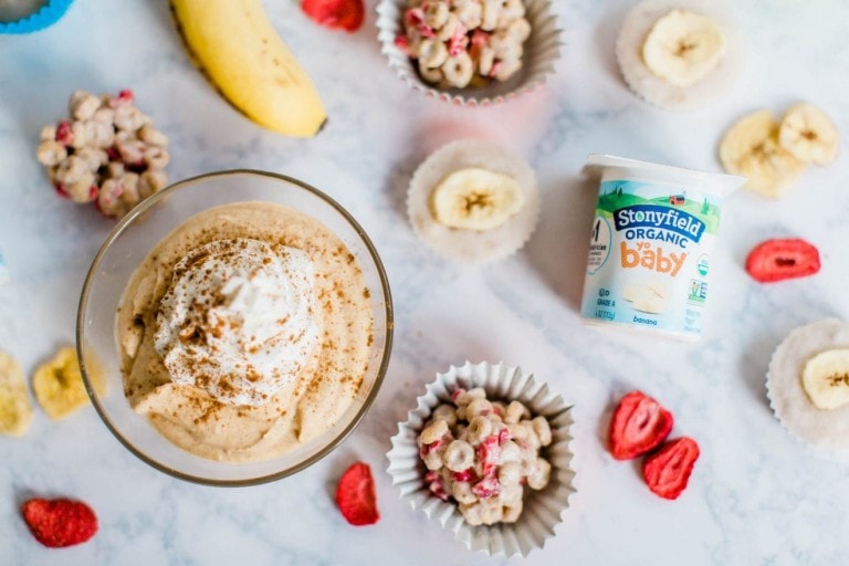 A glass bowl of whipped cream-topped dessert sits on a marble counter surrounded by a yogurt cup labeled "Stonyfield Organic Baby," a banana, freeze-dried fruit pieces, and small cups of cereal with dried strawberries and bananas—delicious inspirations for your yogurt snack recipes.
