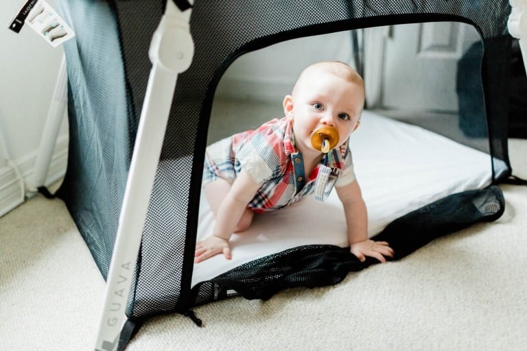 A baby is crawling out of a black and white playpen at grandma's house. The baby is wearing a plaid romper and has a pacifier in their mouth. The playpen is set on a beige carpeted floor with white walls in the background.