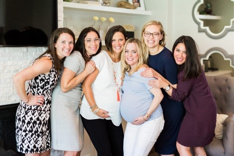 Six women are smiling and standing close together in a well-lit room. Two of the women, visibly pregnant and at the center of the group, are being celebrated in what looks like a blessingway. The room has light-colored walls and modern decor with a large TV on the wall.