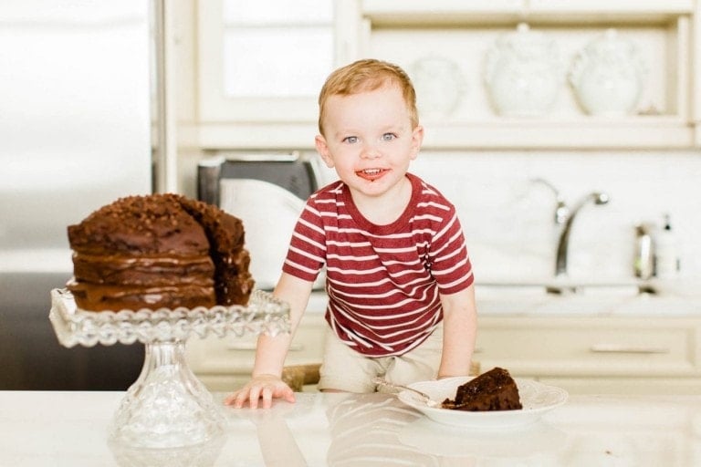 A young child in a red and white striped shirt is standing next to a healthier birthday cake for kids on a glass stand in a bright kitchen. One slice has been cut from the cake and placed on a plate in front of the child. The child is smiling and looking at the camera.