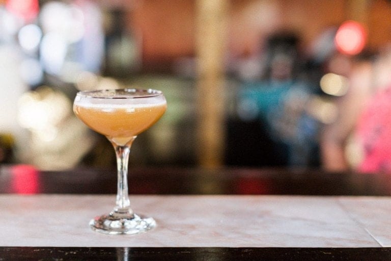 A single cocktail glass filled with a frothy orange mocktail is placed on a marble counter. The background is blurred, showing indistinct shapes and lights that suggest a busy bar or restaurant setting, evoking the cozy vibe of "winter is coming.