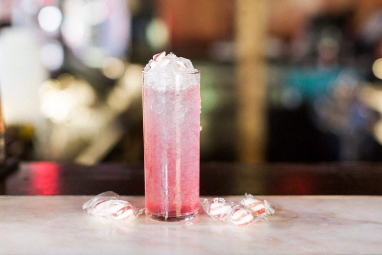A tall glass filled with a pink and clear iced mocktail is placed on a white marble surface. Wrapped peppermint candies are scattered around the base of the glass, evoking a candy land feel. The background is slightly blurred, suggesting an indoor setting.
