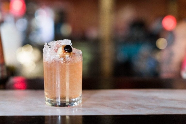 A One Eyed Jack cocktail in a short glass sits on a bar counter. The drink is garnished with a cherry on top of crushed ice. The background is blurred, featuring various colors and lights.