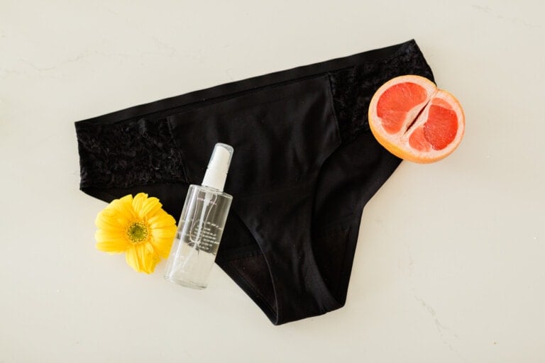 A pair of black women's underwear with lace detail lies elegantly on a white surface. Surrounding it are a yellow flower, a halved grapefruit, and a clear spray bottle, creating a chic display. The scene evokes the delicate beauty of Momotaro peaches in bloom.