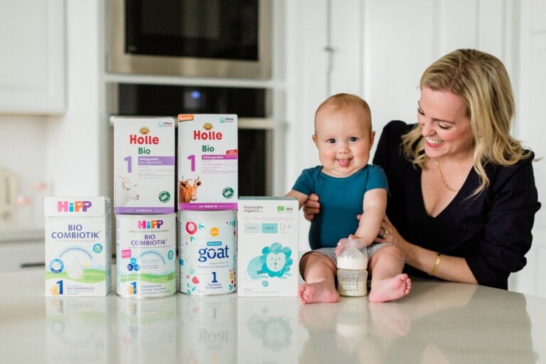 A smiling woman sits next to a baby on a white kitchen countertop. In front of them are several boxes and cans of organic European baby formula from brands like Holle, HiPP, and Kabrita. The baby is holding a bottle of milk. The background features a kitchen stove and cabinets.