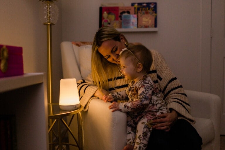A woman sits in a white chair holding a young child on her lap. They are both gazing at a small, lit lamp on a side table. The room is softly lit, and there are children's books on a shelf in the background—essential baby sleep must-haves creating a calm bedtime atmosphere.