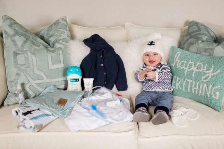 A baby sitting on a couch surrounded by various winter products for baby, including clothes, a bottle, baby lotion, and a knit hat. The couch has decorative pillows, one of which says "happy everything." The baby is wearing a knit hat with an anchor, a patterned sweater, and pants.