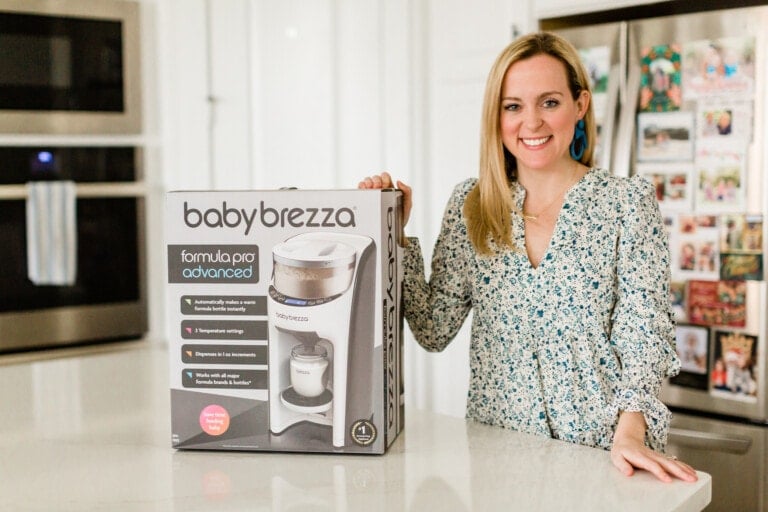 A woman stands in a kitchen next to a Baby Brezza Formula Pro Advanced machine box, smiling warmly in her floral blouse. The kitchen features a stainless steel oven and a fridge adorned with various magnets and photos.