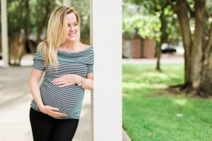 A smiling woman with long blonde hair wearing a black and white striped shirt and black pants is standing and leaning against a white column. She is outdoors, holding her pregnant belly, clearly in her third trimester, in a green, sunlit area.