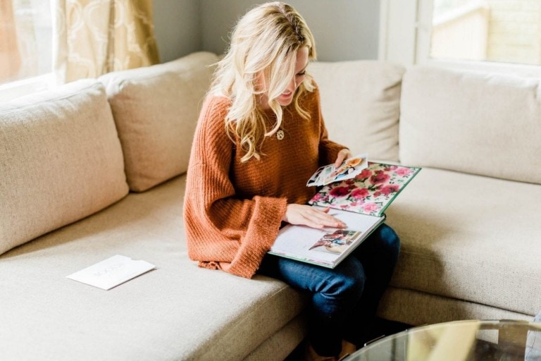 A woman with long blonde hair, wearing a brown knit sweater and jeans, sits on a beige couch looking through a photo album. Beside her is a piece of white paper with writing on it and some recently printed photos from Mootsh. The room has beige curtains and a glass-top table.