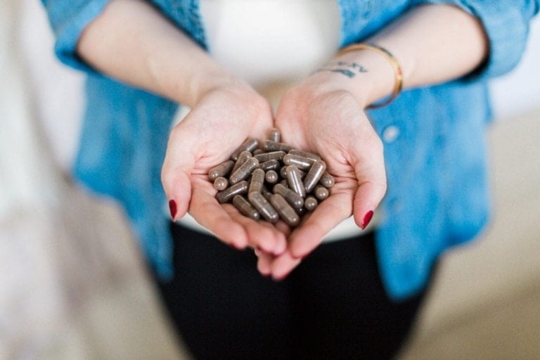 A person with red nail polish holds a handful of dark-colored placenta pills. They are wearing a blue shirt with sleeves rolled up and a white undershirt. A tattoo and bracelets are visible on their left wrist.