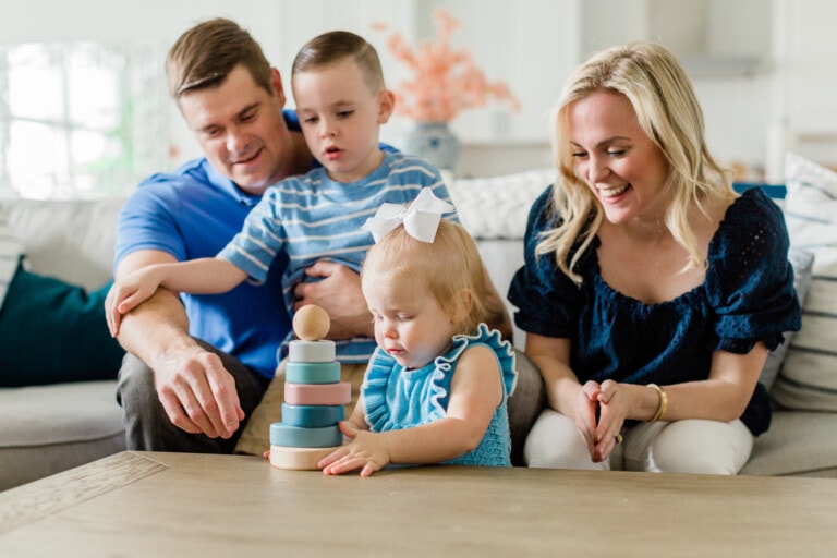 A man and a woman sit on a couch with a boy on the man’s lap and a girl in front of them. They are watching the girl stack colorful rings on a toy tower at a wooden table, enjoying moments of quiet quality time together. They all appear engaged and content.