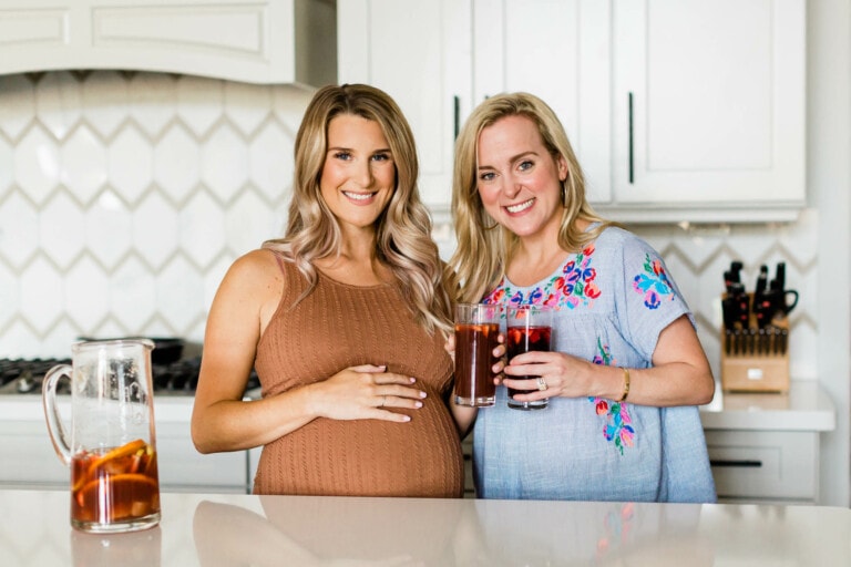 Two women are in a brightly lit kitchen. One is pregnant, wearing a brown dress and touching her stomach. The other wears a light blue dress with floral embroidery and holds up two glasses filled with sangria. A pitcher with the same drink is on the counter.