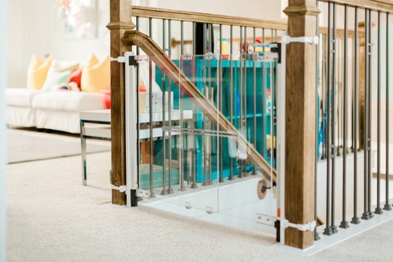 A baby safety gate, crucial for baby proofing, is installed at the top of a staircase in a home. The gate is attached to wooden railings with black metal balusters. A blue bookshelf with various items stands in the background, while a white couch is partially seen to the left.