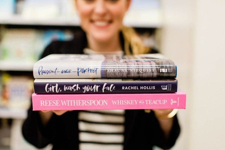 A person holds three books towards the camera, showcasing "Present Over Perfect" by Shauna Niequist, "Girl, Wash Your Face" by Rachel Hollis, and "Whiskey in a Teacup" by Reese Witherspoon—some of the best books for mothers. The person smiles in the background.