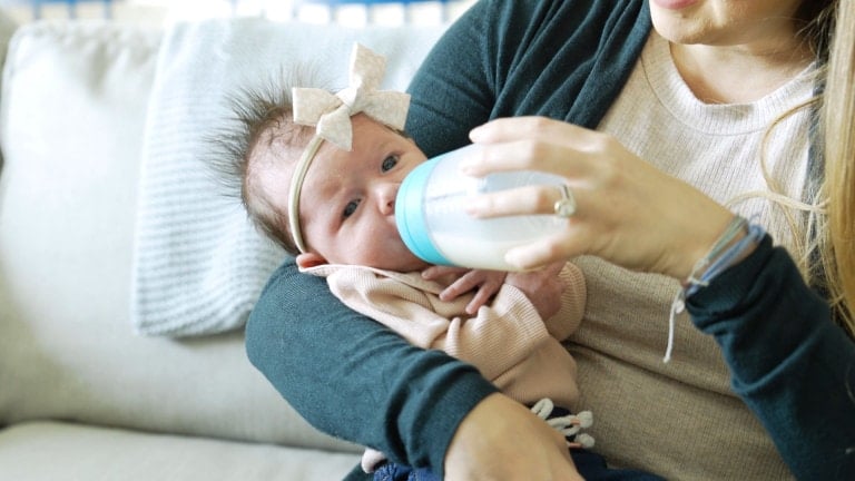 A baby with a bow on their head is being bottle-fed by a woman sitting on a couch. The baby is cradled in the woman's arm, and she is holding the bottle to breastfed baby with her other hand. The woman is wearing a dark sweater and light shirt.