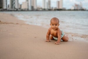 A baby, wearing patterned shorts, crawls on the sandy beach near the shoreline. In the background, there are tall buildings and the ocean under a cloudy sky. Inspired by Hawaiian baby names, this tranquil scene captures both tradition and nature's beauty.