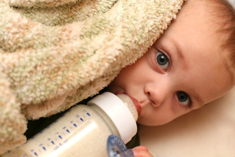 A baby with blue eyes is lying down, partially covered with a blanket. The baby is holding and drinking from a bottle filled with formula. The background is out of focus, drawing attention to the baby's face and the bottle.