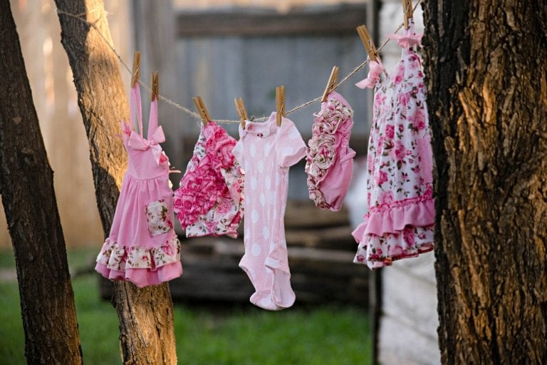 Several small, pink and floral-patterned baby clothes are hanging on a clothesline between two trees, secured with wooden clothespins. The background features a wooden fence and a grass yard, perfect for celebrating a drive-through baby shower.