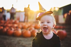 A toddler wearing a small witch hat with a spiderweb design smiles with an open mouth. She is dressed in a black outfit and is sitting outdoors in a pumpkin patch. The background is softly focused with people, pumpkins, and perhaps some spooky girl names echoing the Halloween spirit visible in the sunlight.