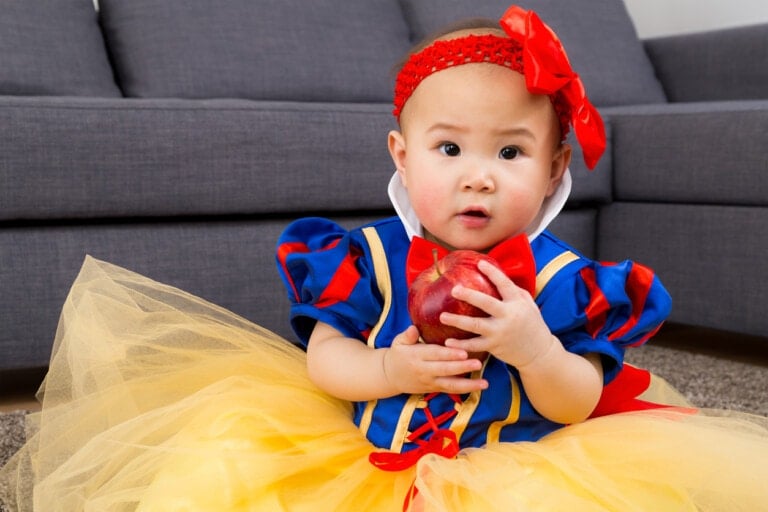 A baby dressed in a Snow White costume sits on the floor holding a red apple. The costume includes a yellow skirt, blue and red bodice, white collar, and a red headband with a bow. The baby looks up with a curious expression, embodying one of those timeless Disney princess names. A gray sofa is in the background.