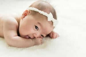 A baby with a white headband featuring a bow is lying on a soft, white surface. The baby is resting their head on their folded arms and has their mouth open, with one fist near their mouth. The scene appears gentle and serene, reminiscent of the calm you feel when picking girl names that start with P.