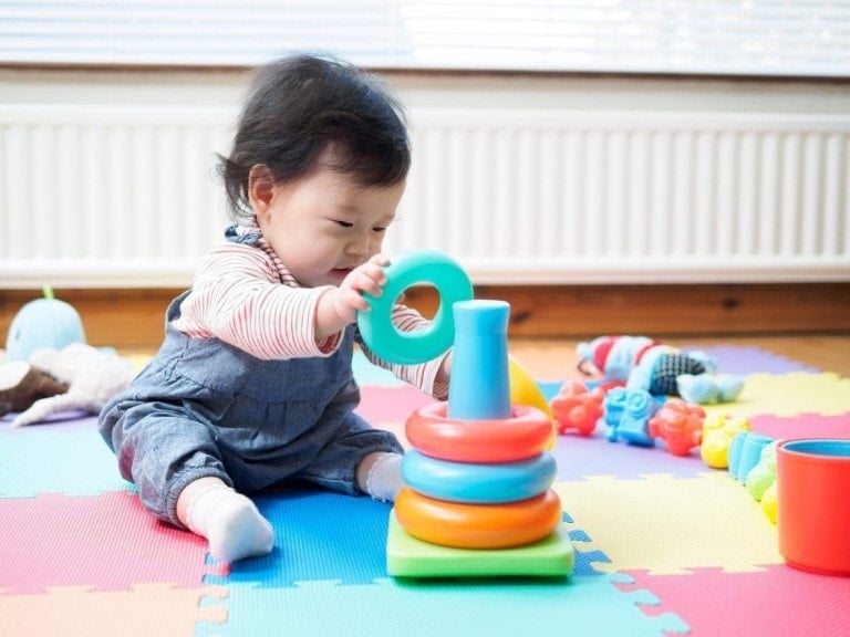 A baby sitting on colorful foam mats is playing with a stack of multicolored rings, marking one of the many exciting milestones in the first year. Other toys are scattered around, including soft plush and plastic toys, with sunlight streaming through window blinds in the background.