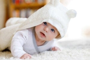 A baby with blue eyes lies on a white carpet, wearing a white hooded outfit with bear ears. The baby gazes curiously at the camera, with the hood partially covering their head. The background is blurred, showing books on a shelf that could inspire e boy names.