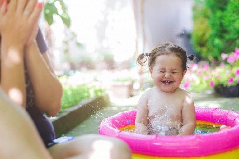 A toddler sits in a pink inflatable pool, splashing water and smiling with eyes closed. An adult, one of the summer must-haves, is next to them, partially visible with hands clapped to their face. The background includes plants and a patio area.