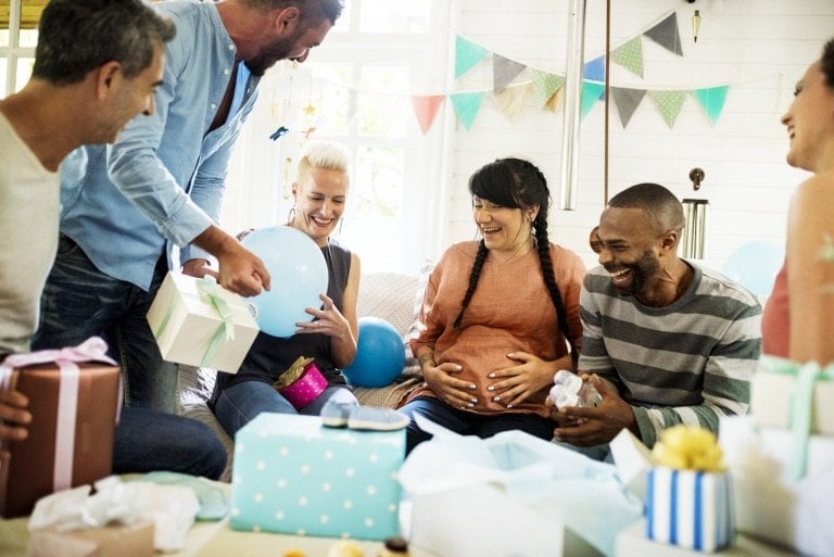 A group of six people are celebrating at a co-ed baby shower. A visibly pregnant woman sits at the center, surrounded by colorful gifts and balloons. The group appears to be indoors in a well-lit room with festive decorations such as bunting.