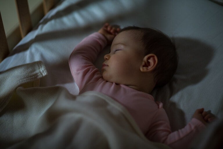 A baby in a pink onesie sleeps on their back in a crib. One arm is raised while the other is covered by a white blanket that also partially covers the baby. The crib's wooden bars are visible in the background. The lighting is dim and soft, leaving parents to wonder when do babies sleep through the night.