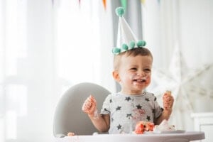A 12-month-old baby wearing a conical birthday hat with pom-poms sits in a high chair, smiling and covered in cake frosting. The background has colorful triangular banners and a star decoration.