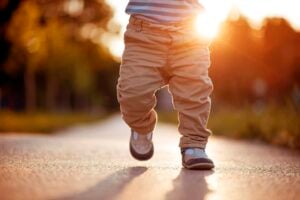 A close-up photo of a 13-month-old reaching milestones while walking outdoors on a paved path. The child is wearing beige pants, a striped shirt, and brown shoes. The sun is setting in the background, illuminating the scene with a warm, golden light.