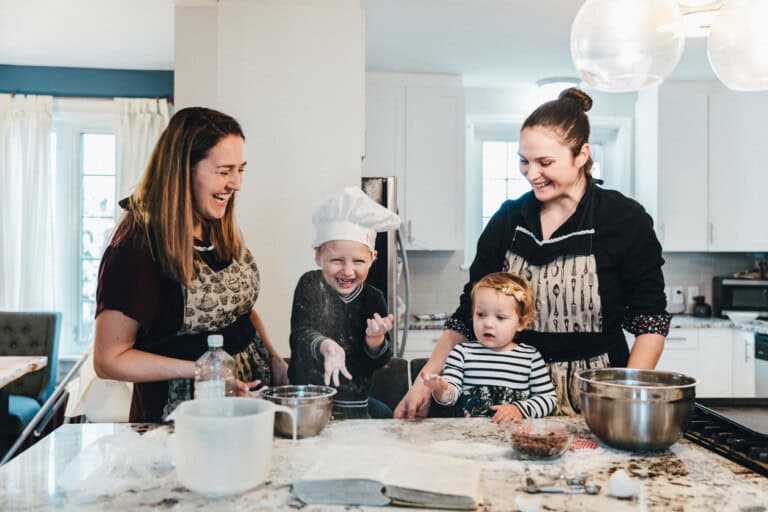 Two women, who appear to be aunts, and two children are baking together in a bright kitchen. One child is wearing a chefs hat. The countertop is covered with baking ingredients and utensils. The group appears happy and engaged in the baking activity.