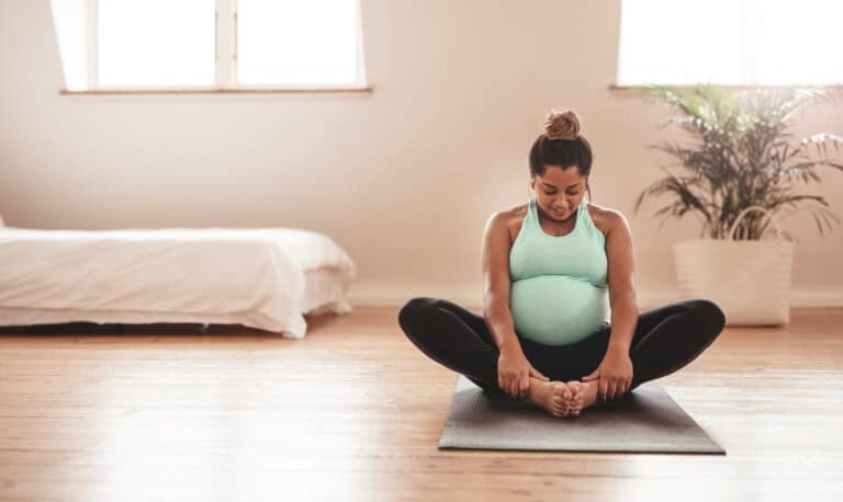 A pregnant woman sits on a yoga mat in a butterfly pose, embracing the benefits of exercise during pregnancy. She wears a turquoise tank top and black leggings in a bright room with wooden floors, a white bed, and a potted plant in the background.