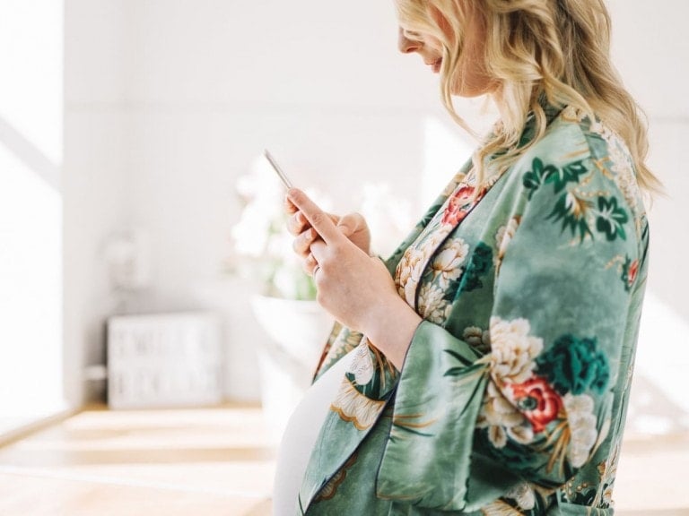 A pregnant woman stands indoors holding a smartphone, presumably browsing the best pregnancy apps. She is wearing a green floral robe and her blonde hair is styled in loose waves. The background is bright and includes a blurred sign and some floral arrangements.