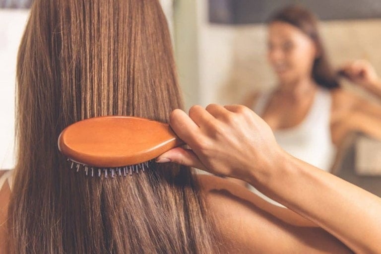 A person with long, straight hair is brushing their hair with a wooden hairbrush in front of a mirror. The focus is on the hand holding the brush while the person's reflection is slightly blurred in the background, pondering whether to dye hair during pregnancy.