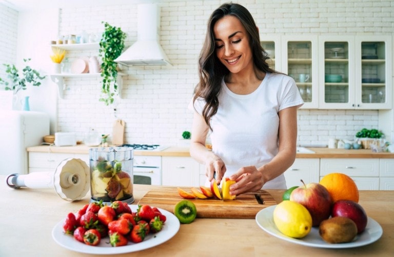 A woman in a white t-shirt is slicing a peach on a wooden cutting board in a bright kitchen. Nearby are plates with a variety of fruits, including strawberries, kiwi, apples, and oranges. A blender filled with chopped fruits sits beside her, ready to create smoothies that boost your immune system.