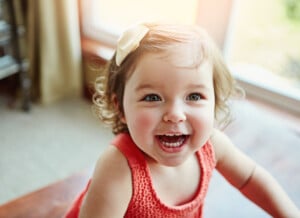 A young child with light brown, curly hair is smiling widely. She is wearing a sleeveless orange top and has a white bow in her hair. Indoors, with sunlight streaming through the window behind her, she looks like she could be one of those adorable girls named Grace or Gia.