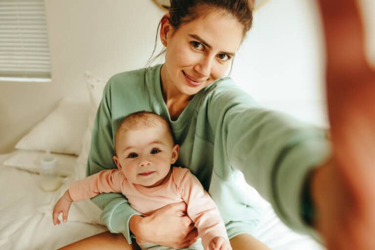 Blogger mom taking a selfie with her baby at home. Millennial mom taking a picture while sitting on the bed with her baby. A woman in a green sweater is sitting on a bed, holding a baby in a pink outfit, both gazing at the camera. In this bright, cozy room with white bedding and pillows.