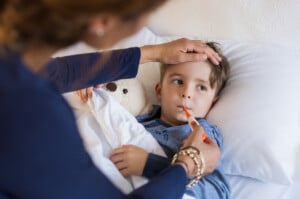 A young child lies in bed, holding a stuffed animal, with a thermometer in their mouth. An adult touches the child's forehead, possibly checking for a fever related to Hand Foot and Mouth Disease. The unwell child looks to the side in a well-lit, cozy bedroom.