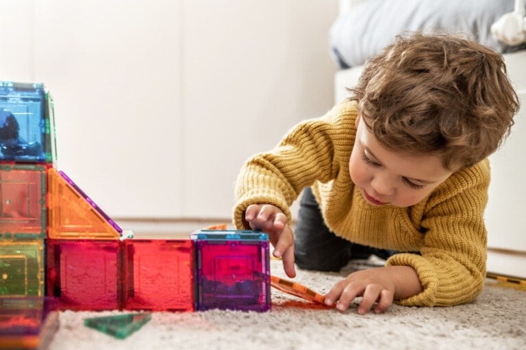 A young child with curly hair, wearing a yellow sweater, is playing with multicolored magnetic tiles on a carpeted floor. The child is focused on assembling the educational toys, forming various shapes and structures. A bed is partially visible in the background.