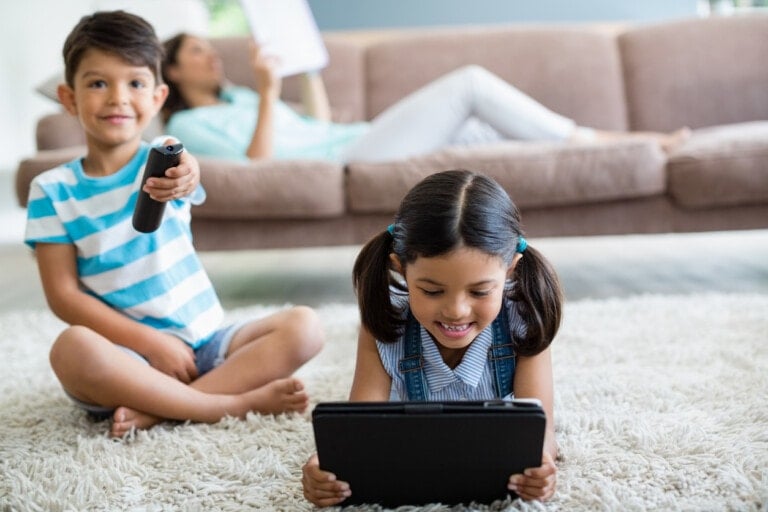 Two children are seated on a carpeted floor in a cozy living room. The boy on the left is holding a TV remote, possibly set to children's shows, and looking at the camera, while the girl on the right is engrossed in her tablet. An adult reclines on the couch in the background, reading a document.