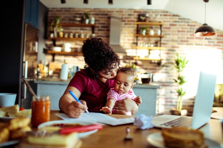 A woman is sitting at a kitchen table with her baby on her lap. She is writing thank-you notes in a notebook, while the baby is touching the pages. The kitchen has an exposed brick wall and shelves with various items. A laptop and breakfast items are on the table.