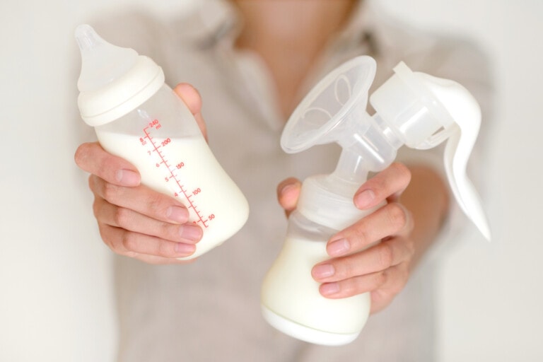 A person is holding a baby bottle filled with milk in their right hand and a manual breast pump with milk in its container in their left hand. The image is focused on the hands and the items, emphasizing how pumping breastmilk is hard work, while the person's face remains out of focus.