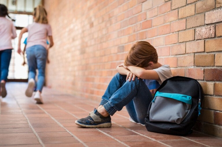A young boy sitting on the floor against a brick wall with his head resting on his arms, his backpack next to him. He seems to be enduring verbal bullying as two children in the background walk away.