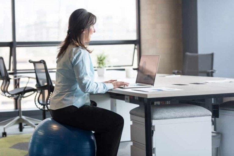 A person is seated on an exercise ball while working on a laptop at a desk in a modern office, embodying the concept of being healthy at work. The individual wears a light blue shirt and black pants. In the background, there are large windows, office chairs, and desks.