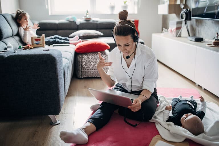 A woman wearing a headset sits on the floor holding a tablet, engaging in a video call. A baby lies on a blanket beside her. In the background, a child sits on a couch, engrossed in a book. The living room, filled with bright natural light, captures the mother-load of multitasking motherhood moments.